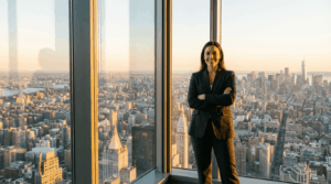 אישה בטוחה בעצמה עומדת ליד חלון משרד המשקיף לקו הרקיע של העיר, יציבה רגועה ומלאת עוצמה, אור יום חמים, צילום תאגידי ריאליסטי, יחס רוחב־גובה 16:9.